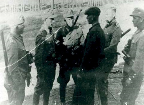 Grupa pięciu wojskowych w niemieckich mundurach z bronią, stojący wokół mężczyzny z cywilnym ubraniu.