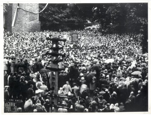 Tłum ludzi. W głębi fragment ceglanej budowli, przy której zwisa dużych rozmiarów, dwubarwna wstęga.