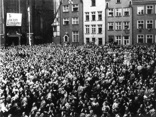 Tłum ludzi na placu. W głębi kamienice oraz fragment kościoła, nad wejściem którego zawieszono planszę z napisem „Te Deum Laudamus”.