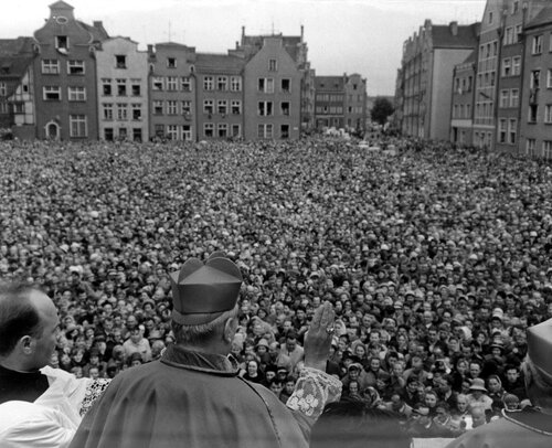 Na pierwszym planie sylwetka prymasa z ręką uniesioną w geście błogosławieństwa nad tłumem ludzi. W głębi kamienice okalające plac.