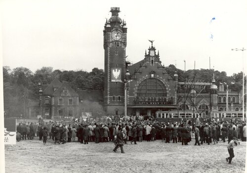 Grupa ludzi wokół metalowej konstrukcji przed budynkiem dworca. W głębi przejeżdżający tramwaj i samochody.