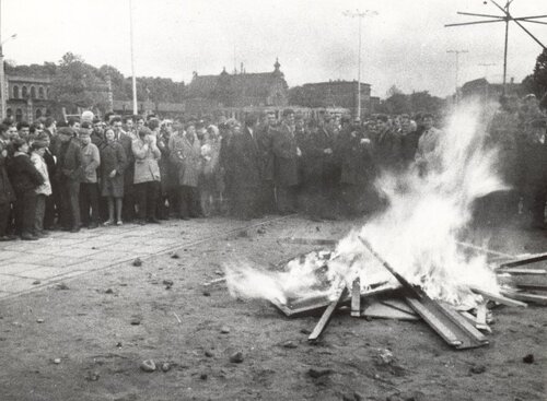 Grupa ludzi skupionych wokół płonącego elementu. W głębi budynek dworca.