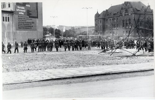 Mundurowi rozstawieni w szerokim szyku przechodzą przez plac, gdzie znajduje się zniszczona konstrukcja. W głębi grupa ludzi, przejeżdżający tramwaj i samochody. W tle budynki zabudowy miejskiej.
