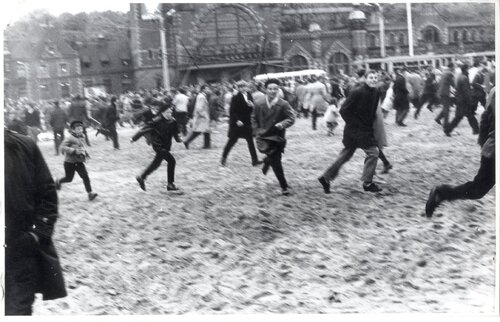 Na pierwszym planie kilka osób w biegu. W głębi grupa ludzi, przejeżdżający tramwaj i samochody. W tle budynek dworca.