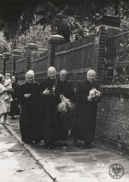 Na pierwszym planie widoczne cztery osoby - duchowni - przechodzące chodnikiem przy murze; w rękach trzymają bukiety kwiatów. Z tyłu siostra zakonna oraz kobieta z dzieckiem na ręku.