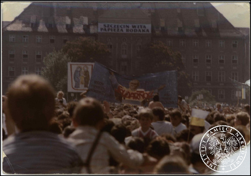 Tłum wiernych na placu. W tle duży budynek. Widoczne transparenty z podobizną Świętej Rodziny oraz Jana Pawła II z napisem „Solidarność”. Na froncie budynku w tle napis: „SZCZECIN WITA PAPIEŻA RODAKA”.