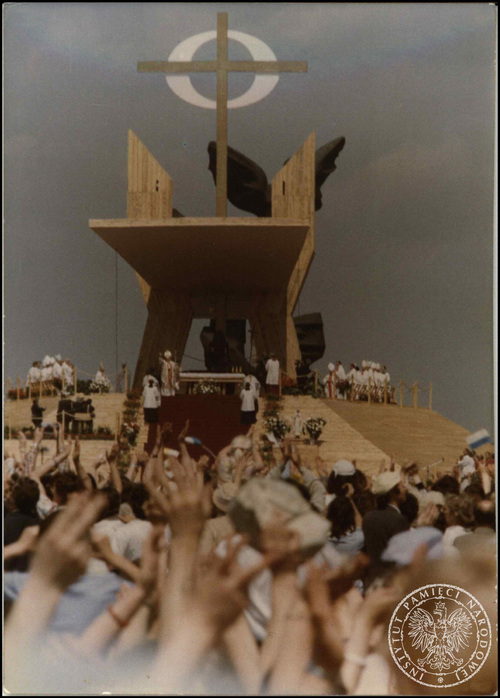 Widok z tłumu wiernych, wznoszących ręce ku górze, na monumentalny, drewniany ołtarz polowy, zwieńczony wysokim, prostym krzyżem. Na ołtarzu, w tym na jego stopniach, stoją osoby duchowne: biskupi, księża oraz - pozdrawiający wiernych - Jan Paweł II.