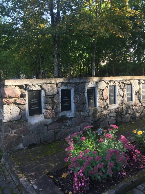 Fragment muru kamiennego z czarnymi tablicami, na których umieszczone są, wyryte białą farbą, imiona i nazwiska (w dwóch wersjach językowych: litewskiej i polskiej) oraz krzyże. Przed murem - rabata kwiatowa, za murem - las.