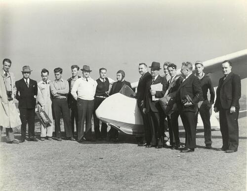 Grupa mężczyzn pozująca do zdjęcia obok szybowca. Jeden z mężczyzn stojący przy kokpicie ma na głowie czapkę pilota.