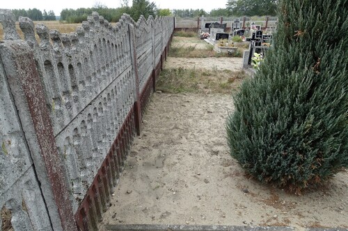 Współczesne zdjęcie przedstawiające fragment terenu cmentarza przy betonowym ogrodzeniu.