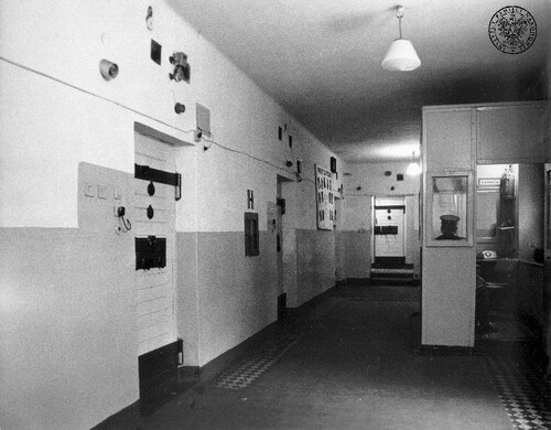 Korytarz więzienny; po lewej stronie drzwi do cel. Nieco w głębi budka strażnicza z widocznym tyłem głowy strażnika.