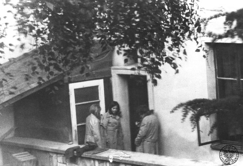Trzy osoby, w tym przynajmniej dwie kobiety, stoją na balkonie, przy otwartych drzwiach balkonowych. Obok - częściowo otwarte okno, nad osobami - pełne liści gałęzie drzewa.