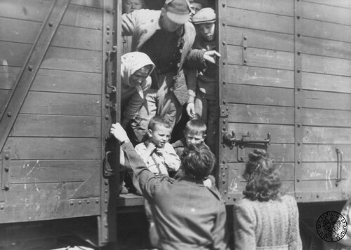 Wagon towarowy; częściowo otwarta ściana wagonu, przez którą wychylają się ludzie: dzieci, kobiety i mężczyźni. Przed wagonem stoją, odwrócone twarzami do ludzi w wagonie, dwie osoby.