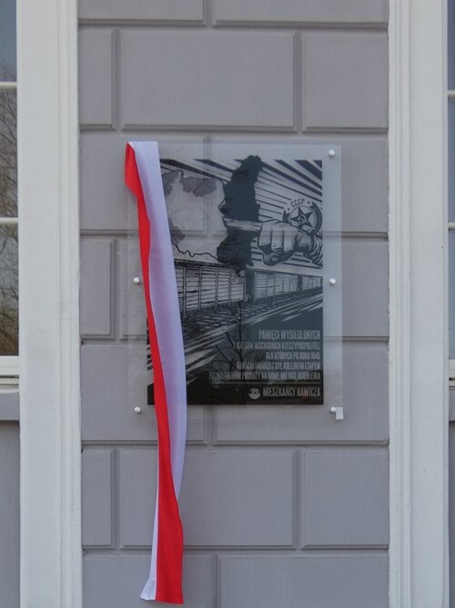 Szklana tablica na murze z jasnych, dużych cegieł. Na tablicy wisi biało-czerwona wstęga.