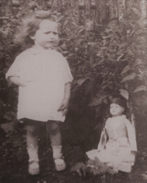 Na archiwalnej fotografii kilkuletnia dziewczynka w białej sukience stoi w ogrodzie. Obok niej lalka w białej sukience.