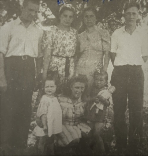 Na archiwalnej fotografii ludzie pozujący do zdjęcia pod drzewem. Stoi dwójka mężczyzn i kobiet. Przed nimi kuca kobieta obejmująca rękoma dwie kilkuletnie dziewczynki.