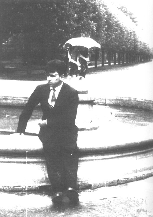 Młody mężczyzna, w garniturze, białej koszuli i z krawatem, siedzi na ozdobnym obmurowaniu fontanny w parku.