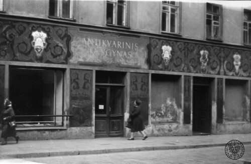 Na szyldzie nad wejściem do antykwariatu napis w języku litewskim „Antikvarinis Knygynas”. Na fasadzie budynku, na wysokości pomiędzy parterem a pierwszym piętrem, namalowano ozdobny wzór. Na chodniku przed budynkiem dwie kobiety, z czego jedna zmierzającą w stronę wejścia do antykwariatu.