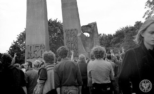 Przed pomnikiem, odwróceni doń twarzami, stoją młodzi ludzie. Z prawej strony widać młodą kobietę, stojącą twarzą do autora zdjęcia. Na pomniku daty: na lewej jego części - „1956”, a na prawej, od dołu - „1968”, „1970”, „1976” i „1980”. Z tyłu pomnika zarośla.