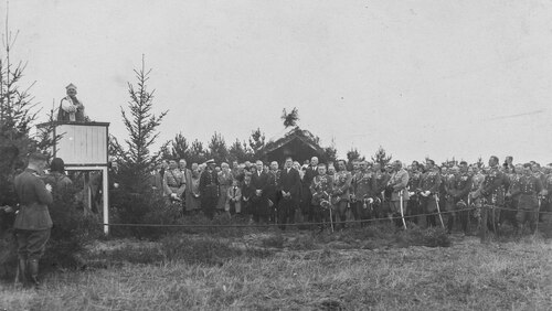 Żołnierze i kilku cywili podczas mszy polowej. Na drewnianym podwyższeniu widoczny przemawiający mężczyzna w stroju hierarchy katolickiego