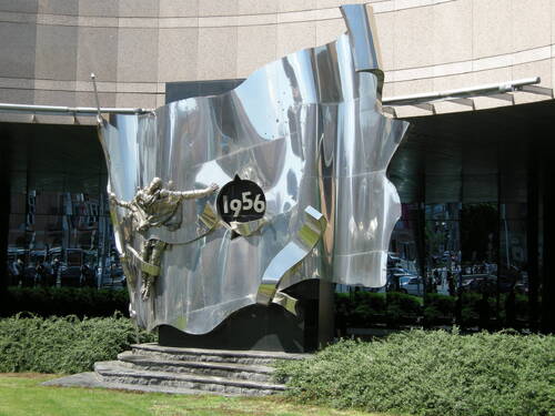 Pomnik z chromowanej stali symbolizujący flagę z otworem w środku, w którym umieszczona jest data: „1956”. Z prawej strony daty na fladze znajduje się rzeźba z wizerunkiem męskiej postaci z rozłożonymi rękoma. Z tyłu nowoczesny budynek z dużą ilością elementów szklanych. Pod pomnikiem schodki i zieleń.