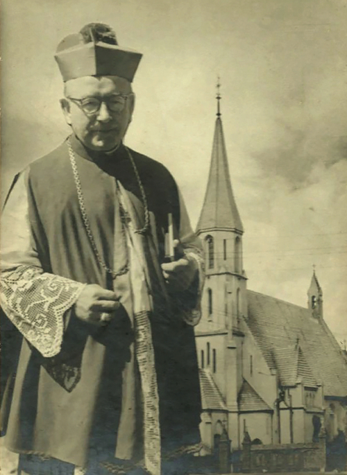 Zdjęcie starszego mężczyzny w stroju katolickiegohierarchy na tle zabytkowego kościoła gotyckiego