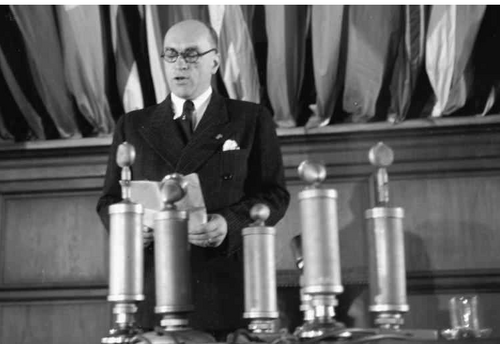 Jarosław Iwaszkiewicz przemawia na Kongresie Pokoju w 1948 roku. Mężczyzna w okularach stoi za pulpitem, na którym umieszczono kilka mikrofonów. Za jego plecami widoczne dolne części flag zatkniętych na drzewcach.