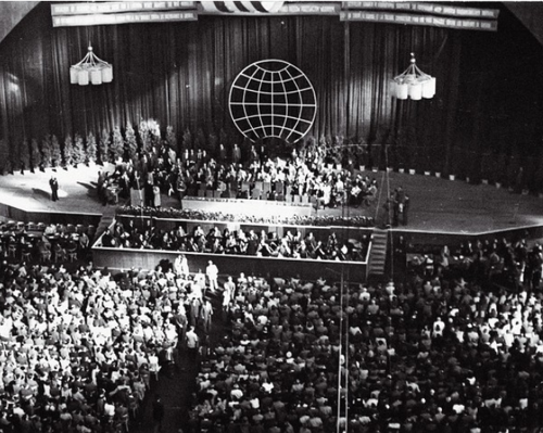 Plenum Kongresu Intelektualistów Wrocław 1948 r. Widok ogólny sali z licznie zgromadzoną publicznością. Duża grupa osób na scenie. W ramach dekoracji na płótnie za sceną graficzne przedstawienie siatki globu ziemskiego.