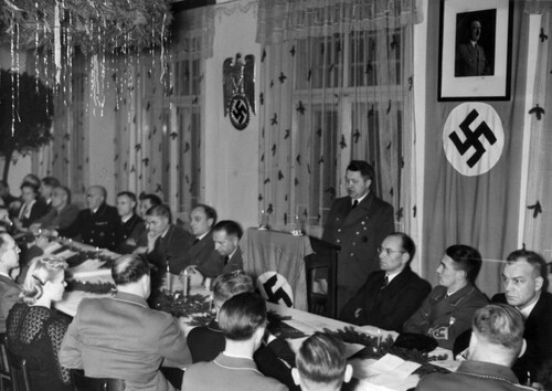 Sala. Długi, świątecznie udekorowany stół, przy którym siedzą ludzie, głównie mężczyźni, ale i parę kobiet. Większość mężczyzn ubrana jest w, jasne lub ciemne, mundury partyjne, część w garnitury. Jeden z mężczyzn przemawia przy trybunie, na której położono ówczesną flagę niemiecką (ze swastyką). Nad stołem z jednej strony wisi portret Hitlera na innej, dużo większej fladze niemieckiej (nazistowskiej), z drugiej nazistowskie godło (orzeł ze złożonymi skrzydłami z wieńcem - w której jest swastyka - w szponach).
