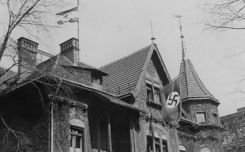 Górna część dużego budynku o złożonym, wieloszczytowym dachu. Ściany budynku są pokryte ozdobną ornamentyką. Na dachu powiewa flaga (niemiecka), a ze szczytowego okna zwisa druga - także niemiecka przy czym w wersji nazistowskiej (ze swastyką).