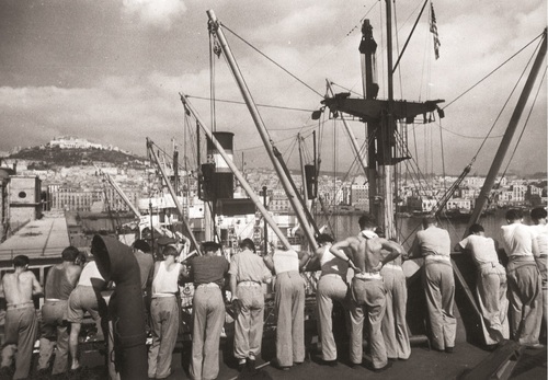 Żołnierze 2. Korpusu przed wyjazdem z Neapolu do Wielkiej Brytanii w 1946 r. Kilkunastu żołnierzy (widoczni tyłem) opierający się o barierę nad kanałem portowym. Widoczne elementy infrastruktury portowej, w tle panorama miasta.