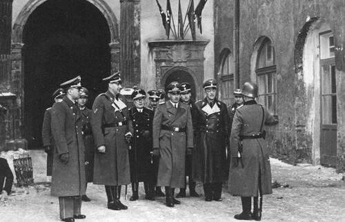 Dziedziniec zamkowy, wokół mury zamkowe, w tle brama. Grupa mężczyzn ubranych w niemieckie, zimowe ubrania służbowe: płaszcze, wysokie, czarne buty i czapki oficerskie lub urzędnicze. Gubernator Hans Frank przyjmuje meldunek od niemieckiego żołnierza, który na zdjęciu stoi tyłem i ma na głowie hełm. Na murze osadzonych kilka niemieckich flag nazistowskich.