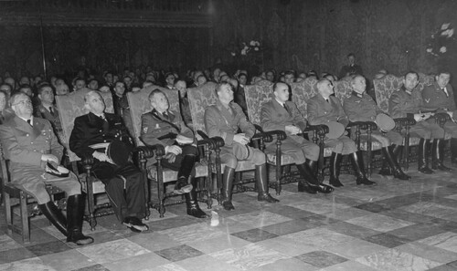 Duże pomieszczenie z błyszczącą posadzką. W kilku rzędach siedzi audytorium złożone z niemieckich wojskowych lub urzędników. Mężczyźni w pierwszym rzędzie siedzą na ozdobnych, wysokich fotelach. Mają mundury z emblematami nazistowskimi i wysokie, czarne buty, w rękach trzymają czapki. Wszyscy są gładko ogoleni i przyjmują wyrazy twarzy osób poważnych, wypełniających bardzo ważne zadania.