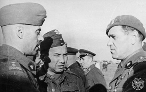 Na pierwszym planie trzech rozmawiających mężczyzn w polskich mundurach oficerskich, dwóch ma na głowie furażerki, jeden - beret. W tle inne osoby, w tym dwóch mężczyzn w mundurach sowieckich.