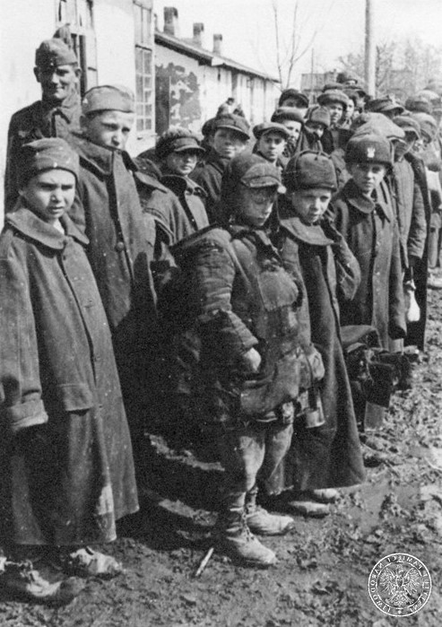 Błotnisty plac, z tyłu zabudowania. Dzieci ubrane są w łachmany - zniszczoną odzież, płaszcze wojskowe, czapki i furażerki wojskowe różnych wzorów (polskie i sowieckie). Z tyłu jeden z żołnierzy polskich.