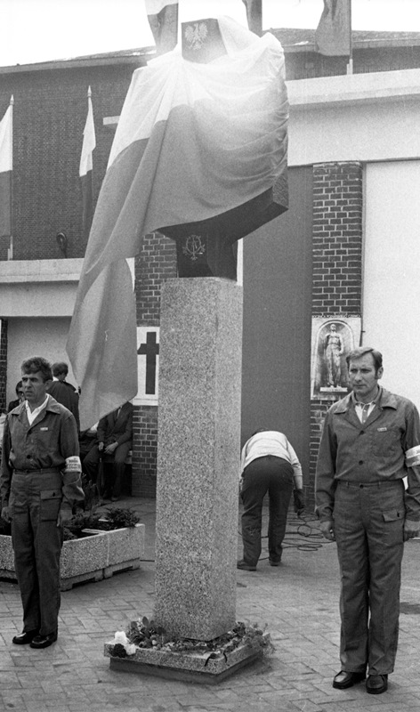 Ceremonia odsłonięcia Czerwcowej tablicy pamiątkowej przed bramą główną HCP, 27 czerwca 1981 r. Tablica wieńcząca cokół osłonięta biało-czerwoną flagą, znad której wystaje element z godłem PRL.