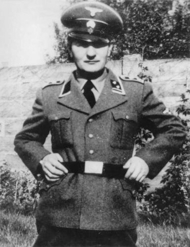 Pieter Menten pozujący do zdjęcia w mundurze SS, 1941 r. W tle betonowe ogrodzenie i roślinność.