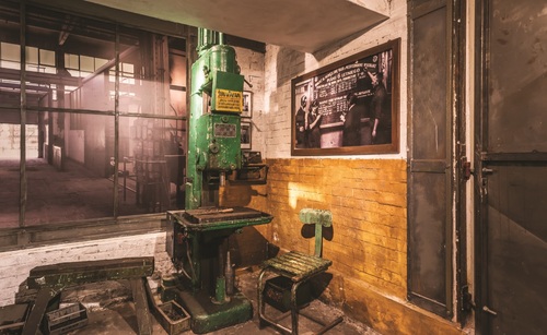 Fragment ekspozycji przedstawiający miejsce pracy w Zakładach Przemysłu Metalowego im. Józefa Stalina w Poznaniu. Zaaranżowana przestrzeń z elementami ceglanej ściany, urządzeniem fabrycznym oraz archiwalnymi zdjęciami, z których jedno w dużym rozmiarze imituje przestrzeń hali fabrycznej.