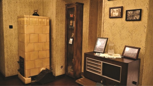 Rekonstrukcja mieszkania rodziny poznańskiej z lat pięćdziesiątych XX w. Przestrzeń z piecem kaflowym, radiem w drewnianej zabudowie, wazon, serwetka, zdjęcia rodzinne, witryna z bibelotami.