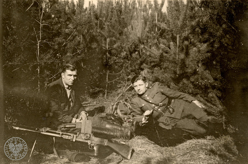 Zagajnik, niskie drzewa, między innymi jodły. Na trawie, po prawej stronie zdjęcia, leży mężczyzna w mundurze, z przypiętym do pasa granatem. Z lewej strony zdjęcia siedzi drugi mężczyzna, ubrany po cywilnemu. Przed nim, na trawie, znajduje się ręczny karabin maszynowy.