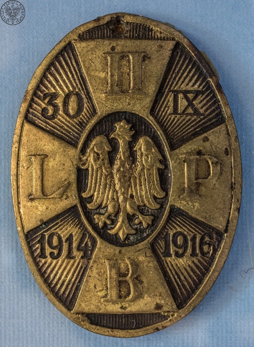 Owalna odznaka pamiątkowa II Brygady Legionów Polskich z blachy mosiężnej przedstawiająca krzyż, na którego ramionach widnieją wypukłe monogramy: „II”, „B[rygada]”, „L[egionów]”, „P[olskich]”. W środku krzyża umiejscowiony orzeł jagielloński. W polach, pomiędzy ramionami krzyża, na tle rozchodzących się promieni, wyryto: „30”, „IX”, „1914” i „1916”.