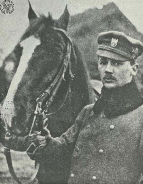 Młody mężczyzna w wojskowych płaszczu i czapce, trzymający za uzdę konia.
