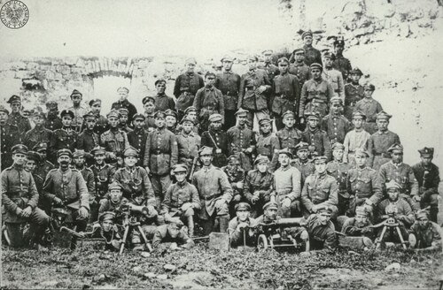 Duża grupa żołnierzy - młodych mężczyzn - w kilku rzędach stojących, półklęczących i leżących. Żołnierze są ubrani w kurtki wojskowe i różnego typu czapki, między innymi maciejówki. Przed pierwszym rzędem żołnierzy, leżących w trawie, stoją trzy ciężkie karabiny maszynowe z taśmami z amunicją i skrzynkami amunicyjnymi. W tle ruiny jakiejś budowli.