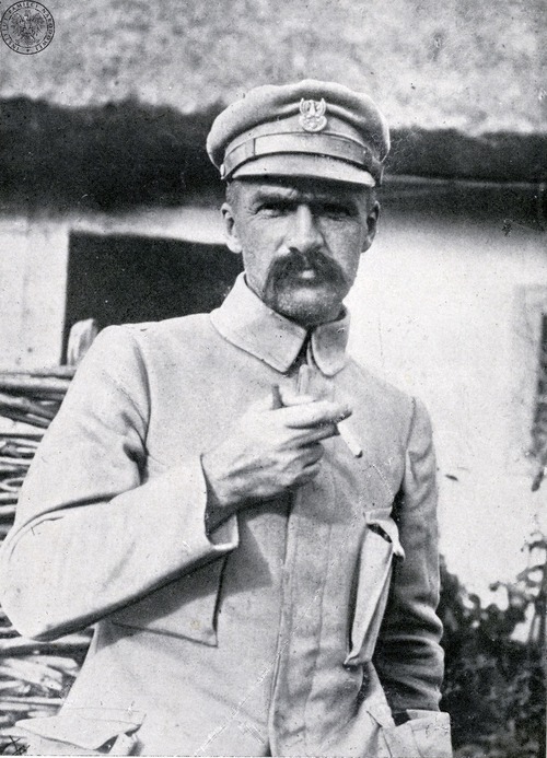 Józef Piłsudski - mężczyzna w średnim wieku z dużymi wąsami, trzymający w prawej dłoni papierosa - w mundurze legionowym bez dystynkcji, w maciejówce na głowie. W tle fragment budynku (być może wiejskiej chaty).