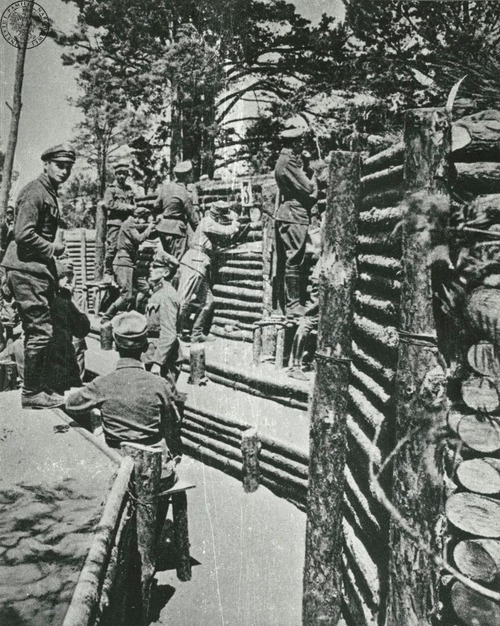 Grupa żołnierzy w parupoziomowym okopie ziemnym zabezpieczonym ściankami z pali drewnianych. Paru żołnierzy obserwuje przedpole, dwóch innych mierzy z karabinów przez otwory strzelnicze. Nad okopem rosną wysokie drzewa.