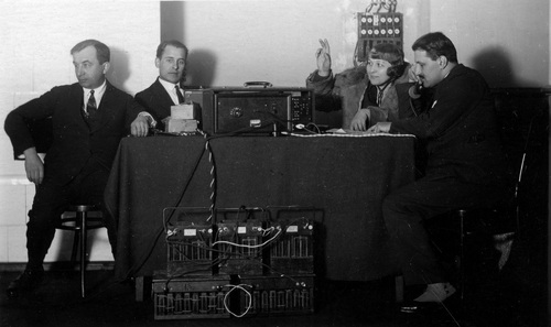 Witold Hulewicz w towarzystwie innych pracowników rozgłośni Polskiego Radia w Wilnie, 1929 r. Trójka mężczyzn i kobieta pozują do zdjęcia, przyjmując różne pozy przy stole, na którym rozłożono aparaturę radiową.