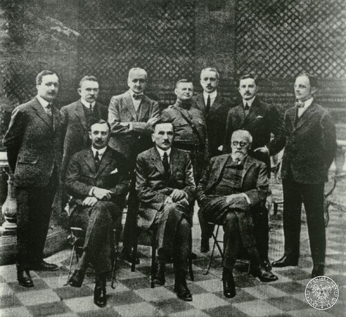 Pozowane zdjęcie grupowe. Z przodu, na krzesłach, siedzi trzech mężczyzn. Za nimi stoi siedmiu innych mężczyzn. Oprócz jednego z mężczyzn, ubranego w mundur, pozostali mają na sobie garnitury z charakterystycznymi w ówczesnym okresie (początek XX w.) prostokątnymi, zaokrąglonymi na końcach kołnierzykami u koszul.