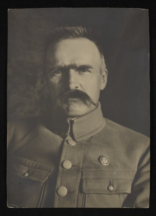 Józef Piłsudski - zdjęcie do połowy piersi. Mężczyzna w średnim wieku z dużymi wąsami, o ostrym, przenikliwym wzroku, krzaczastych brwiach, krótko ostrzyżonych, nieco nastroszonych włosach. Ma na sobie prostą kurtkę wojskową.