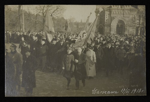 Miasto. Ulicą lub placem przechodzą w dużej liczbie uczestnicy manifestacji patriotycznej, niosąc sztandary. Maszerują osoby w różnym wieku, mężczyźni i kobiety. Starsi ubrani są elegancko, zgodnie z ówcześnie panującą modą (np. mężczyźni mają na głowie meloniki). Na czele maszerujących na zdjęciu wyróżnia się dwóch młodych chłopców, sądząc po charakterystycznych nakryciach głowy - prawdopodobnie gimnazjalistów. W tle dawna zabudowa miejska oraz drzewa.