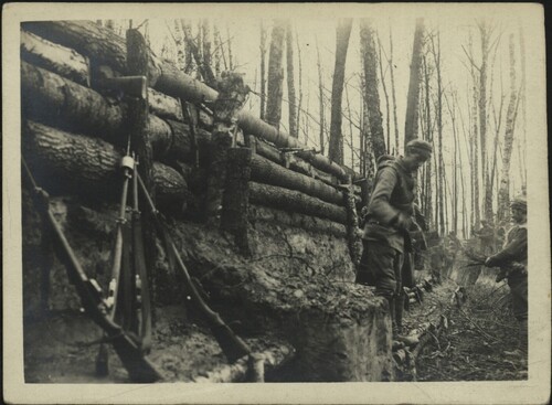 Leśny okop, w którym stoi grupa żołnierzy umundurowanych zgodnie z wzorem legionistów polskich w armii austro-węgierskiej podczas I wojny światowej. Widoczne również, ustawione w tzw. koźle, karabiny piechoty. Okop wzmocniony jest rzędami drewnianych pali.